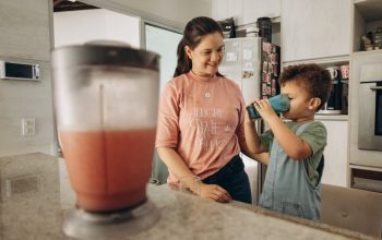 A mother smiles as her son drinks a smoothie from a blue cup in the kitchen, conveying family bonding.